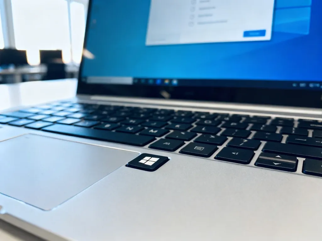 Klawiatura nowoczesnego laptopa z widocznym klawiszem Windows oraz rozmyte menu ustawień systemu w tle.